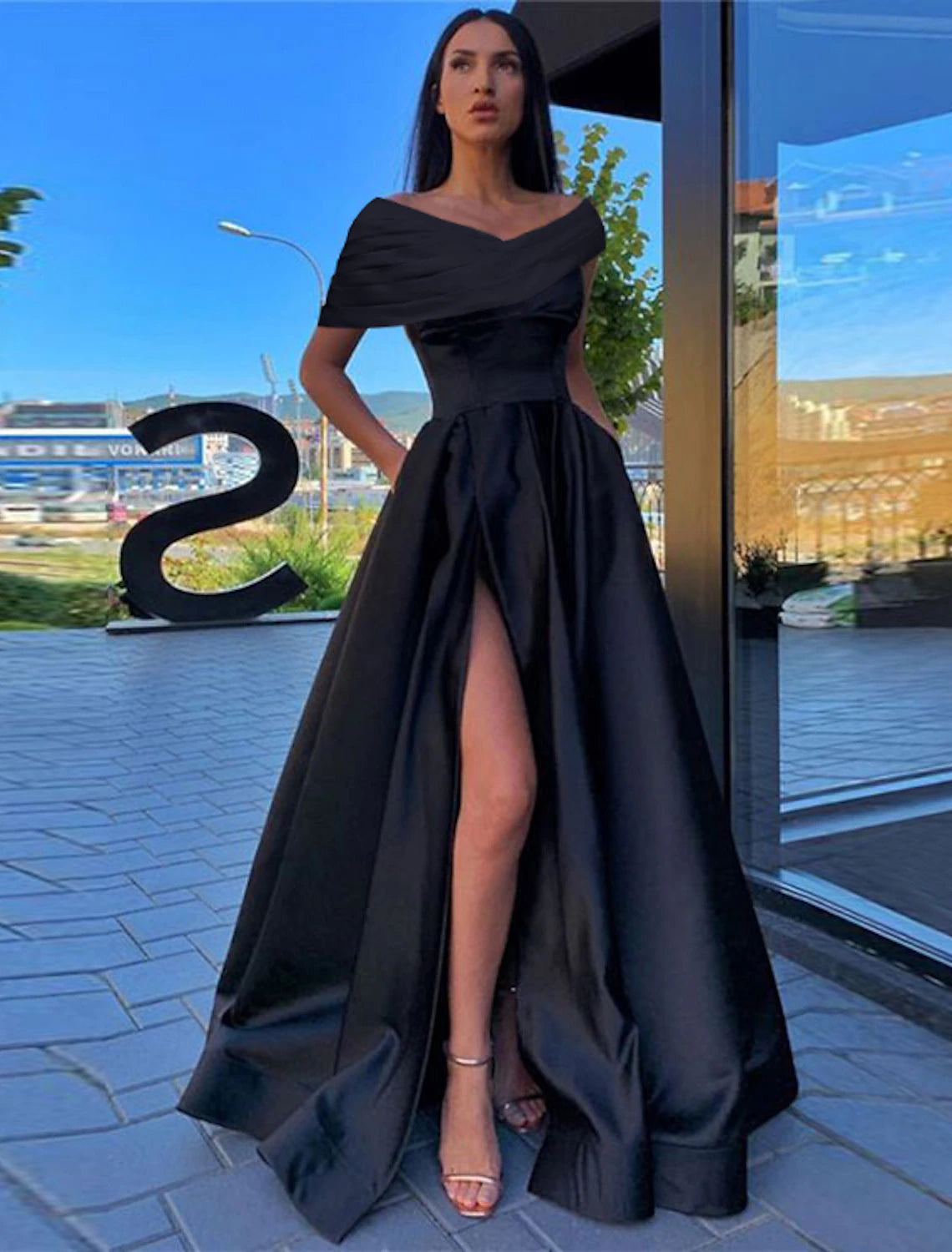 A woman wearing a black A-line off-shoulder short-sleeve satin dress with a ruched slit, posing elegantly.